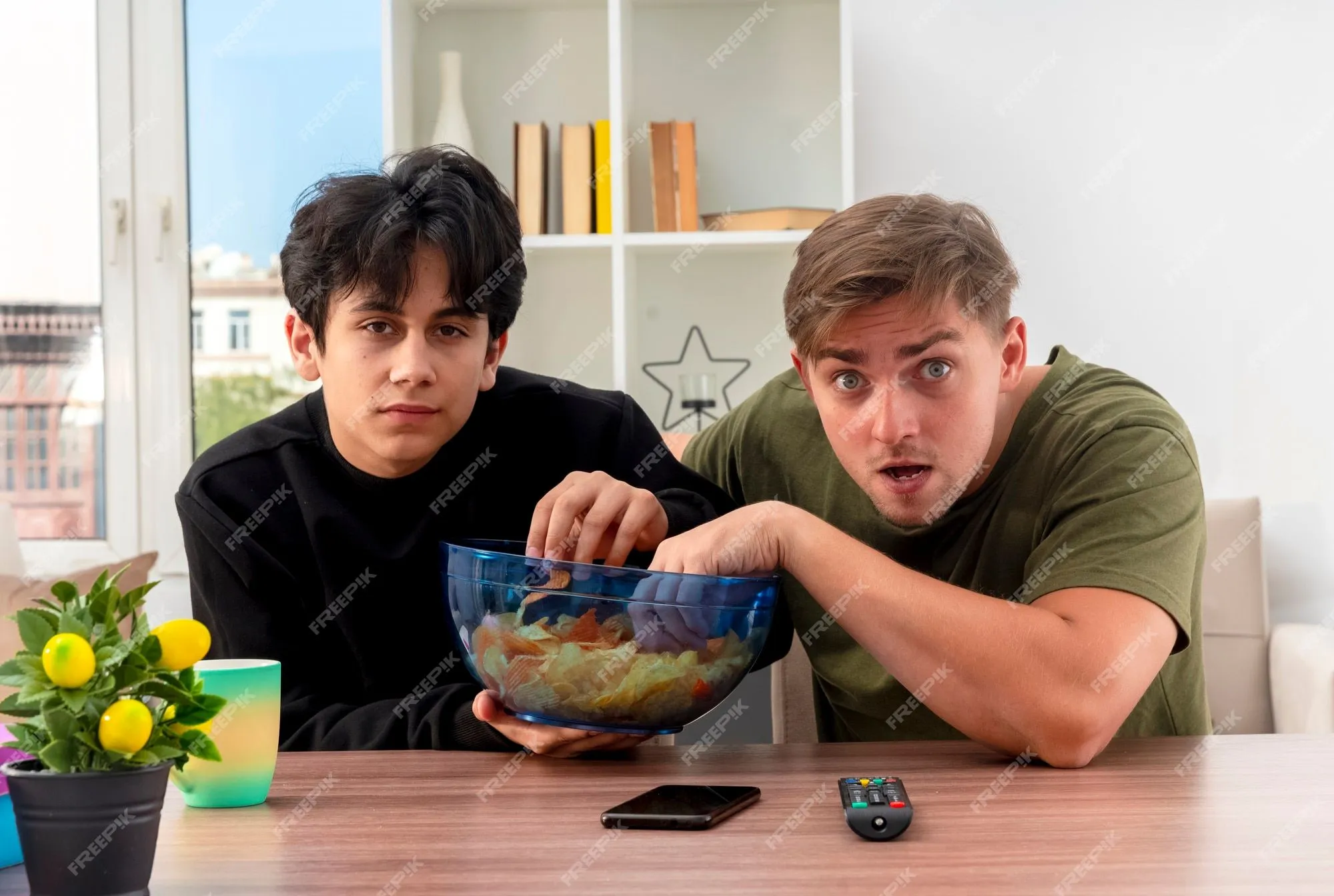 Ansiosos jóvenes rubios y morenos guapos se sientan en la mesa sosteniendo y comiendo un tazón de patatas fritas dentro de la sala | Foto Gratis Ansiosos jóvenes rubios y morenos guapos se sientan en la mesa sosteniendo y comiendo un tazón de patatas fritas dentro de la sala | Foto Gratis