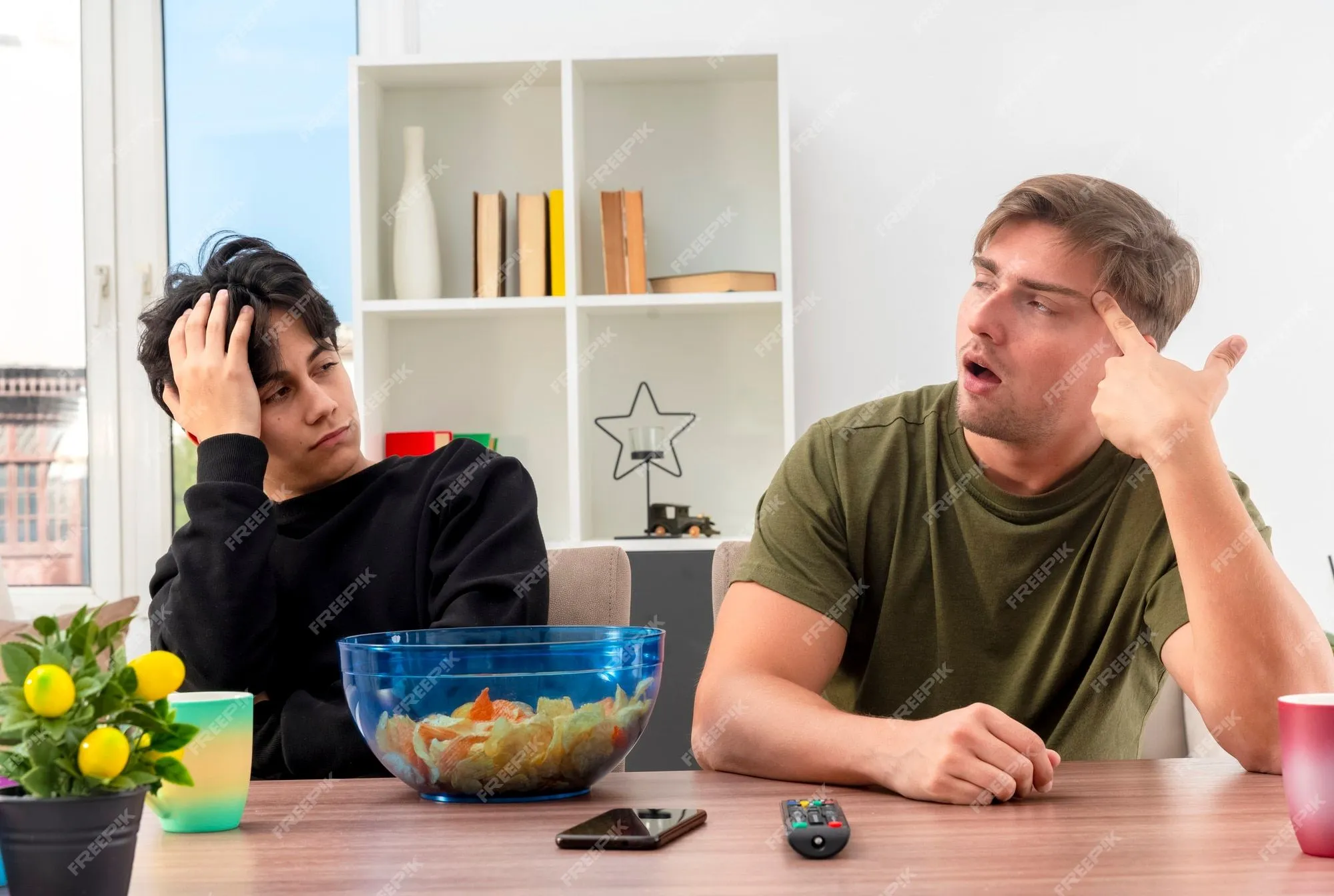 Chicos guapos rubios y morenos jóvenes molestos se sientan a la mesa con un cuenco de chips, teléfono y control remoto de tv poniendo la mano en la cabeza mirando el uno al otro | Foto Gratis Chicos guapos rubios y morenos jóvenes molestos se sientan a la mesa con un cuenco de chips, teléfono y control remoto de tv poniendo la mano en la cabeza mirando el uno al otro | Foto Gratis
